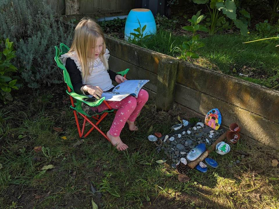 A child sits in a green folding chair and reads next to a fairy garden.