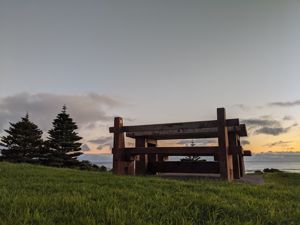 A wooden picnic table on grass in the foreground is shot to resemble a piece of modern art. The sun sets over the sea in the background. Two large trees are at the left of the image.