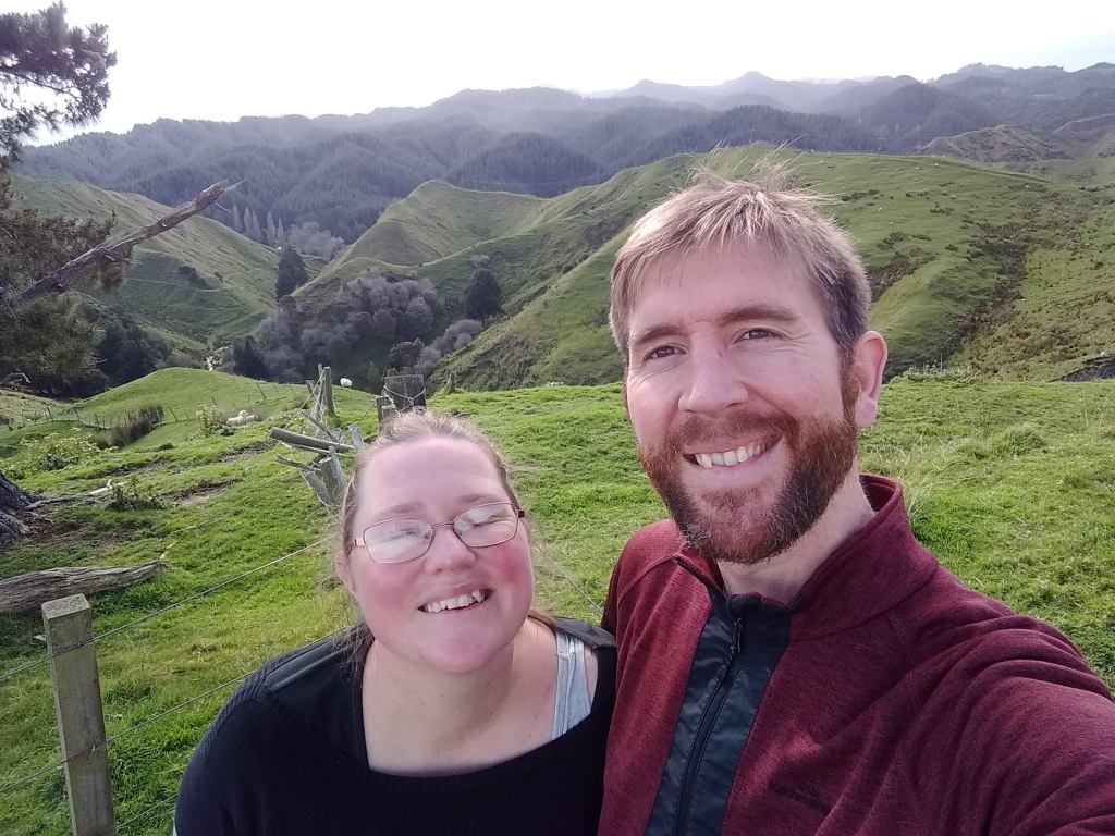 A woman and a man smile in the foreground, with a view of pointed green hills of farmland behind.